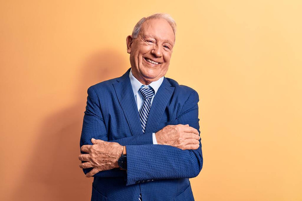 Senior handsome grey-haired businessman wearing elegant suit over yellow background happy face smiling with crossed arms looking at the camera. Positive person. - Photo, Image