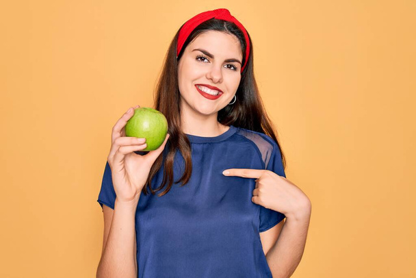 Nuori kaunis tyttö syö tuoretta orgaanista terveellistä vihreää omenaa keltaisella taustalla yllätyksellä kasvot osoittaen sormella itseään - Valokuva, kuva