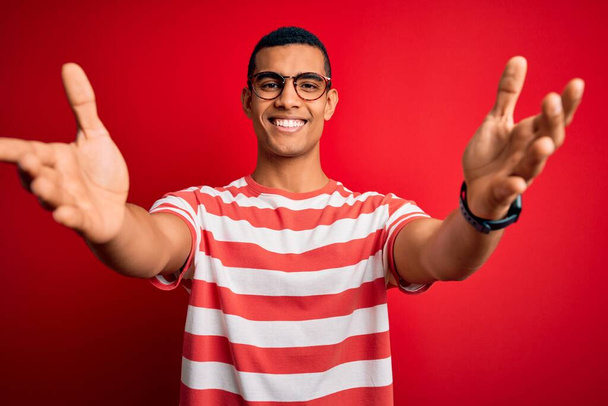 Junger gutaussehender afrikanisch-amerikanischer Mann in lässigem gestreiftem T-Shirt und Brille, der lächelnd mit offenen Armen in die Kamera blickt, um sich zu umarmen. Fröhlicher Ausdruck des Glücks. - Foto, Bild