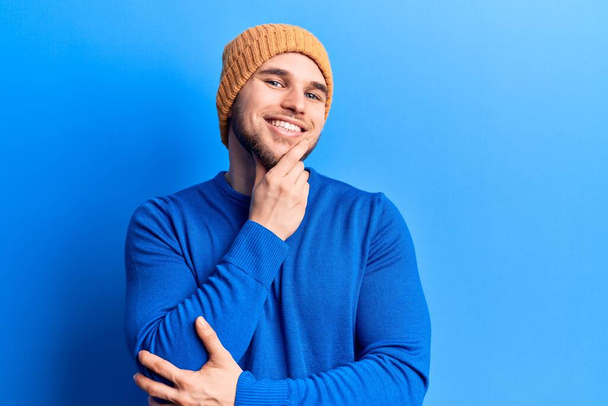Ein junger gutaussehender Mann in lässigem Pullover und Wollmütze lächelt selbstbewusst in die Kamera, mit verschränkten Armen und der Hand am Kinn. Positives Denken.  - Foto, Bild