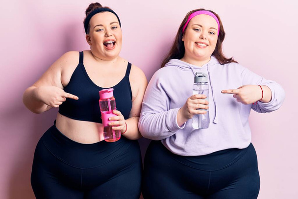 Young plus size twins wearing sportswear drinking bottle of water smiling happy pointing with hand and finger  - Foto, Bild