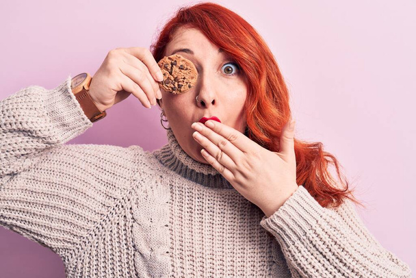 Mladá krásná zrzavá žena stojící s čokoládovou sušenkou přes oči zakrývající ústa rukou, šokovaná a vystrašená z omylu. Překvapený výraz - Fotografie, Obrázek
