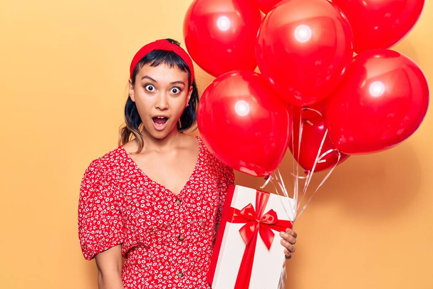 Young woman holding balloons with gift scared and amazed with open mouth for surprise, disbelief face  - Photo, Image