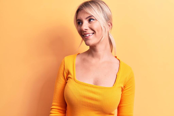 Νεαρή όμορφη ξανθιά γυναίκα φορώντας casual t-shirt στέκεται πάνω από απομονωμένο κίτρινο φόντο κοιτάζοντας προς τα πλάγια, να χαλαρώσετε προφίλ ποζάρουν με φυσικό πρόσωπο και αυτοπεποίθηση χαμόγελο. - Φωτογραφία, εικόνα