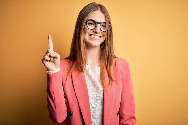 Young beautiful redhead woman wearing jacket and glasses over isolated yellow background showing and pointing up with finger number one while smiling confident and happy. - Photo, image