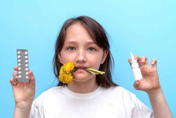 Retrato de una niña con flores en los dientes y pastillas antialérgicas y spray en las manos sobre un fondo azul. El concepto de alergia estacional al polen de las flores
. - Foto, Imagen