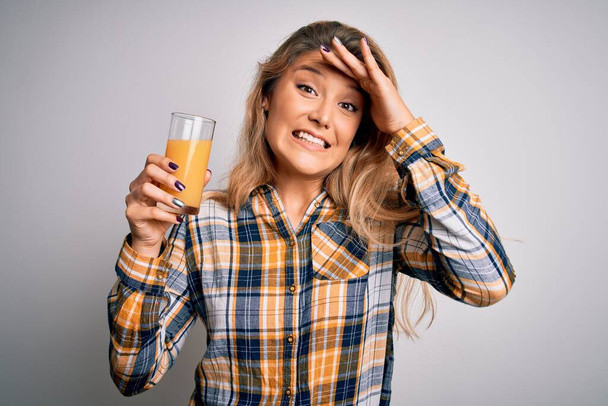 Young beautiful blonde woman drinking glass of healthy orange juice over white background stressed with hand on head, shocked with shame and surprise face, angry and frustrated. Fear and upset for mistake. - Photo, image