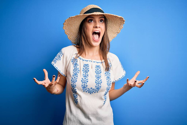 Young beautiful brunette woman wearing casual t-shirt and summer hat over blue background crazy and mad shouting and yelling with aggressive expression and arms raised. Frustration concept. - Photo, Image