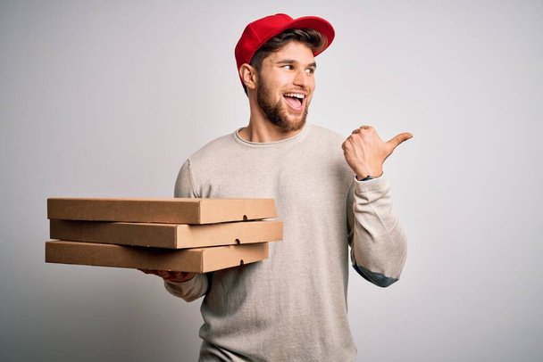 Young blond delivery man with beard and blue eyes holding cardboards of pizza pointing and showing with thumb up to the side with happy face smiling - Photo, Image
