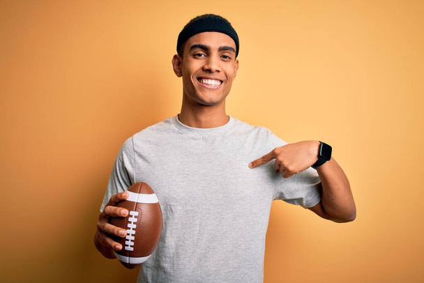 Fiatal jóképű afro-amerikai férfi tartja rögbi labda felett elszigetelt sárga háttér meglepetés arc ujjal mutogatott magára - Fotó, kép