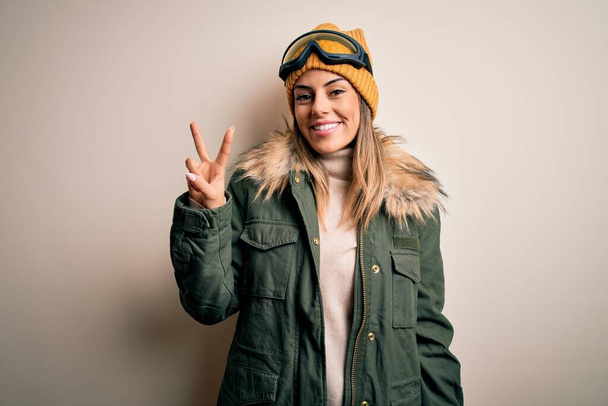 Young brunette skier woman wearing snow clothes and ski goggles over white background showing and pointing up with fingers number two while smiling confident and happy. - 写真・画像