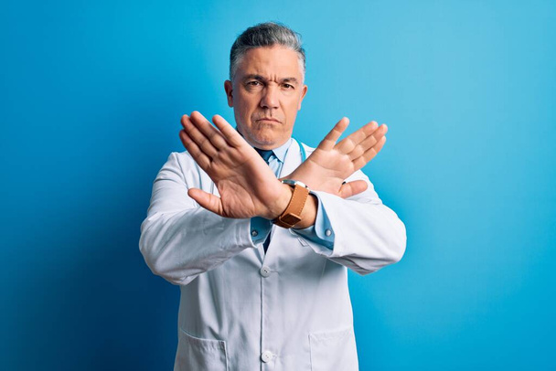 Μεσήλικας όμορφος γκριζομάλλης γιατρός με παλτό και μπλε στηθοσκόπιο έκφραση απόρριψης σταυρωτά χέρια κάνοντας αρνητικό πρόσημο, θυμωμένο πρόσωπο - Φωτογραφία, εικόνα