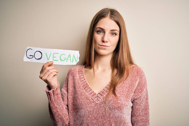 Junge schöne rothaarige Frau, die nach vegetarischem Essen fragt, hält Papier mit veganer Botschaft mit selbstbewusstem Gesichtsausdruck und ernstem Denken in der Hand - Foto, Bild