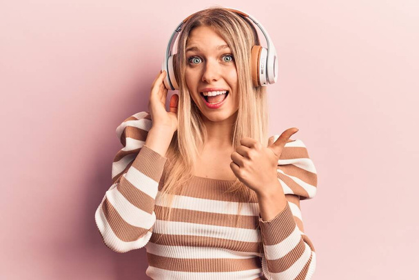 Νεαρή ξανθιά γυναίκα που ακούει μουσική χρησιμοποιώντας ακουστικά δείχνοντας τον αντίχειρα προς τα πλάγια χαμογελώντας χαρούμενη με ανοιχτό στόμα  - Φωτογραφία, εικόνα