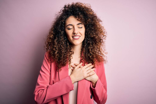 Young beautiful businesswoman with curly hair and piercing wearing elegant jacket smiling with hands on chest with closed eyes and grateful gesture on face. Health concept. - Fotoğraf, Görsel
