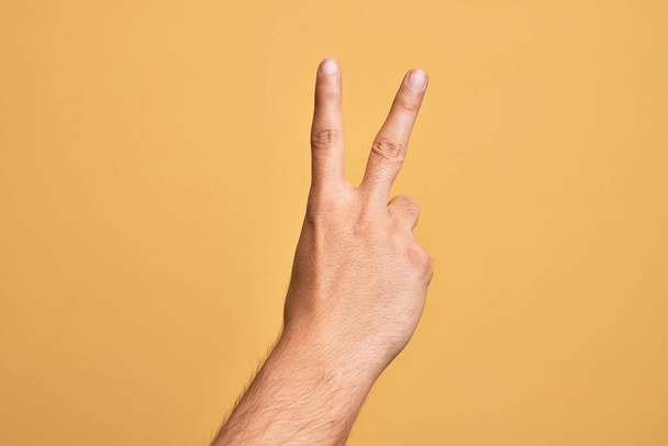 Mão de jovem caucasiano mostrando dedos sobre fundo amarelo isolado contando número 2 mostrando dois dedos, gesticulando vitória e símbolo vencedor - Foto, Imagem