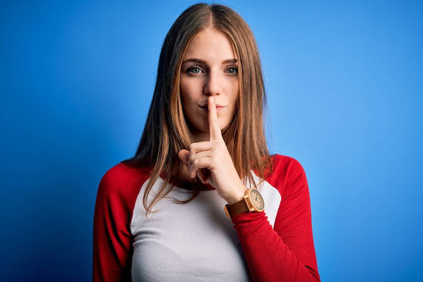 若い美しい赤毛の女性は、孤立した青の背景の上にカジュアルなTシャツを着て唇に指で静かにするように求めている。沈黙と秘密の概念. - 写真・画像