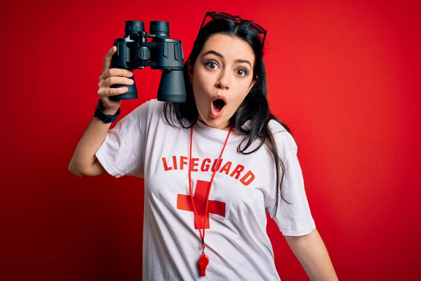 Young lifeguard woman wearing secury guard equipent holding binoculars over red background scared in shock with a surprise face, afraid and excited with fear expression - Photo, Image