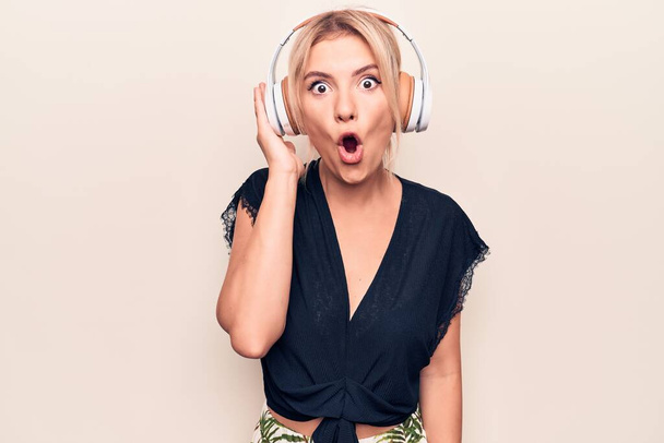 Young beautiful blonde woman listening to music using headphones over white background scared and amazed with open mouth for surprise, disbelief face - Fotó, kép
