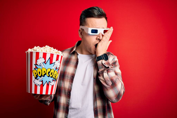Young handsome caucasian man wearing 3d movie glasses and eating popcorn Yawning tired covering half face, eye and mouth with hand. Face hurts in pain. - Photo, Image