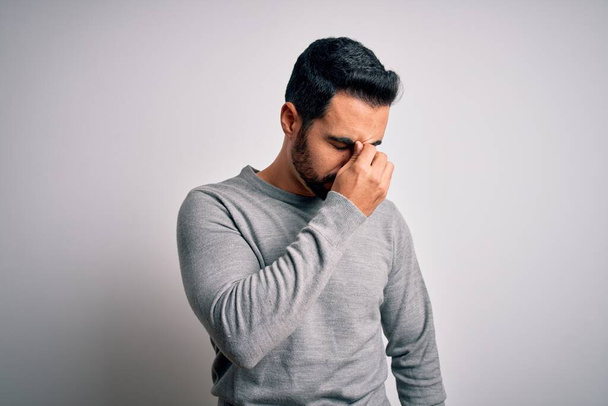 Young handsome man with beard wearing casual sweater standing over white background tired rubbing nose and eyes feeling fatigue and headache. Stress and frustration concept. - Foto, immagini