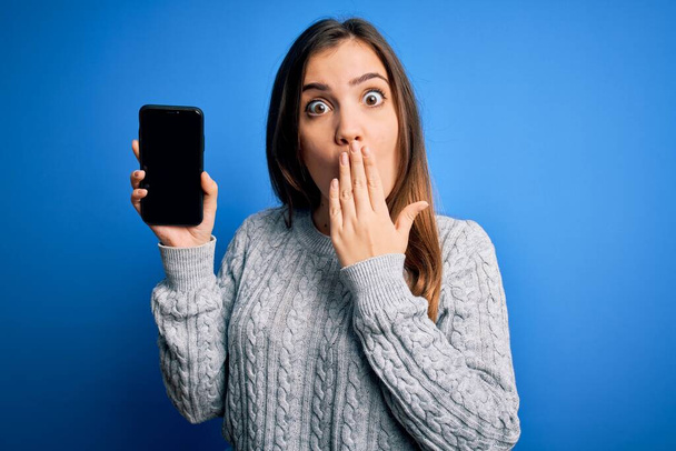 Mladá žena ukazuje prázdný smartphone obrazovku přes modré izolované pozadí kryt ústa s rukou šokované hanbou za omyl, výraz strachu, strach v tichu, tajné koncepce - Fotografie, Obrázek