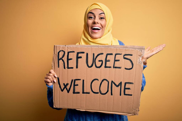 Hermosa mujer árabe con hiyab islámico sosteniendo pancarta con los refugiados mensaje de bienvenida muy feliz y emocionado, expresión ganadora celebrando la victoria gritando con una gran sonrisa y las manos levantadas
 - Foto, imagen