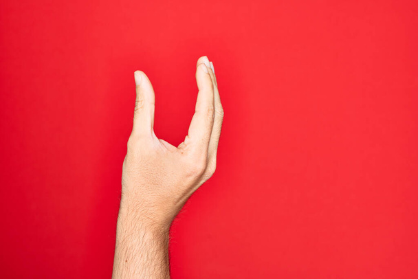 Käsi valkoihoinen nuori mies näyttää sormet yli eristetty punainen tausta poiminta ja ottaen näkymätön asia, pitämällä esine sormin osoittaa tilaa - Valokuva, kuva