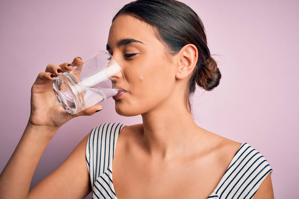 Young beautiful brunette woman drinking glass of healthy water to refreshment standing over isolated pink background - Photo, Image