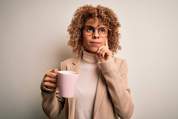 Jonge Afrikaanse amerikaanse krullende vrouw drinken mok koffie over geïsoleerde witte achtergrond serieus gezicht denken over vraag, zeer verward idee - Foto, afbeelding