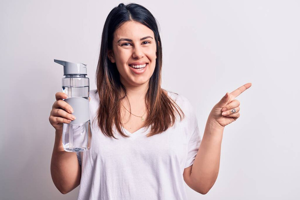 Молодая красивая брюнетка женщина пьет бутылку воды на изолированном белом фоне улыбаясь счастливо указывая рукой и пальцем в сторону
 - Фото, изображение