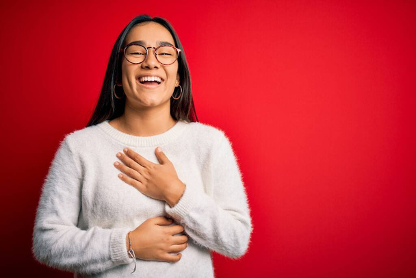 Mladá krásná asijská žena na sobě ležérní svetr a brýle přes červené pozadí s úsměvem a hlasitě se smát, protože legrační šílený vtip s rukama na těle. - Fotografie, Obrázek