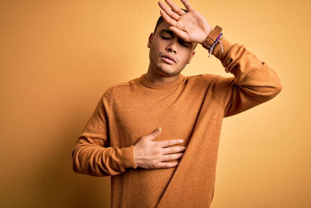Młody Brazylijczyk noszący luźny sweter stojący nad odizolowanym żółtym tłem Dotykający czoła z powodu choroby i gorączki, grypy i przeziębienia, choroby wirusowej - Zdjęcie, obraz