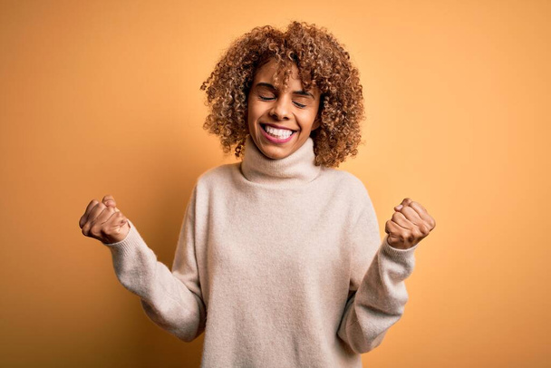 Giovane bella donna africana americana indossa maglione dolcevita su sfondo giallo molto felice ed eccitato facendo gesto vincitore con le braccia alzate, sorridente e urlando per il successo. Concetto di celebrazione. - Foto, immagini