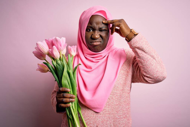 Young african american plus size woman wearing muslim hijab holding bouquet of pink tulips worried and stressed about a problem with hand on forehead, nervous and anxious for crisis - Foto, Bild