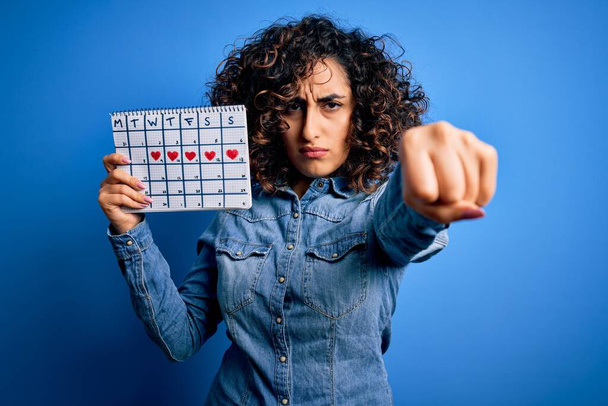 Junge schöne, lockige arabische Frau mit Zeitrahmen-Kalender, der den Menstruationszyklus kontrolliert, genervt und frustriert schreit vor Wut, verrückt und schreit mit erhobener Hand, Wut-Konzept - Foto, Bild