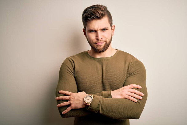 Young blond man with beard and blue eyes wearing green sweater over white background skeptic and nervous, disapproving expression on face with crossed arms. Negative person. - Photo, Image