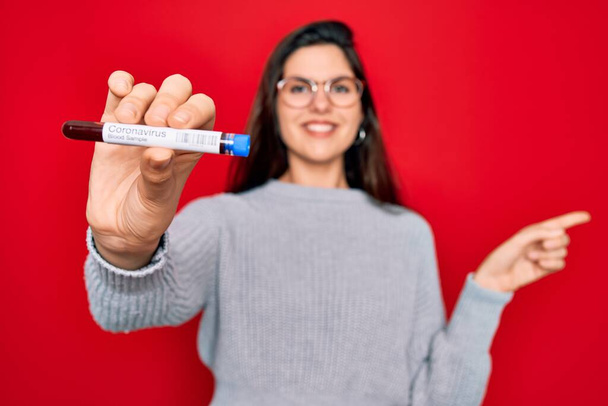 Young beautiful girl wearing glasses holding laboratory blood sample of coronavirus disease very happy pointing with hand and finger to the side - Fotoğraf, Görsel