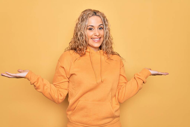 Junge schöne blonde, sportliche Frau in lässigem Sweatshirt vor gelbem Hintergrund, lächelnd mit beiden Händen geöffnete Handflächen, Präsentation und Werbung Vergleich und Balance - Foto, Bild