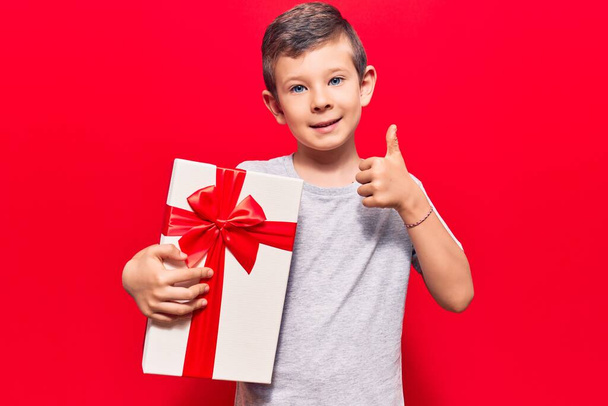 Roztomilé blonďaté dítě držící dárek šťastný a pozitivní úsměv, palec nahoru dělá vynikající a schvalovací znamení  - Fotografie, Obrázek