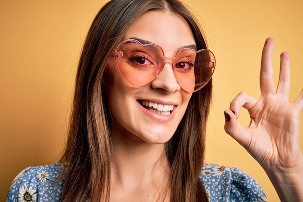 Young beautiful brunette woman wearing funny sunglasses with heart over yellow background doing ok sign with fingers, excellent symbol - Photo, Image
