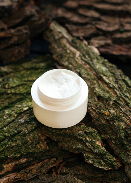 Cosmetic cream jar on tree bark piece side view. Organic cosmetology botanical concept. White plastic container with natural moisturizing balm and greenery. Female skin care accessory. - Photo, Image