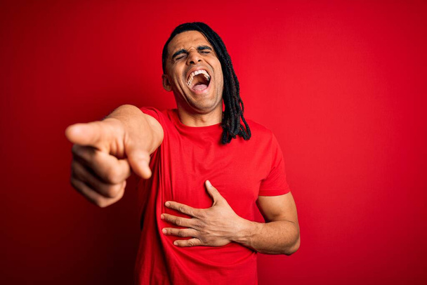 Young handsome african american afro man with dreadlocks wearing red casual t-shirt laughing at you, pointing finger to the camera with hand over body, shame expression - Fotó, kép