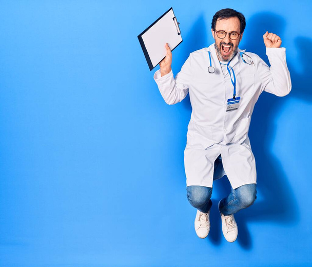 Keski-ikäinen komea lääkäri mies yllään stetoskooppi ja takki hymyillen onnellinen. Hyppy kasvoilla pitäen leikepöydällä yli eristetty sininen tausta - Valokuva, kuva