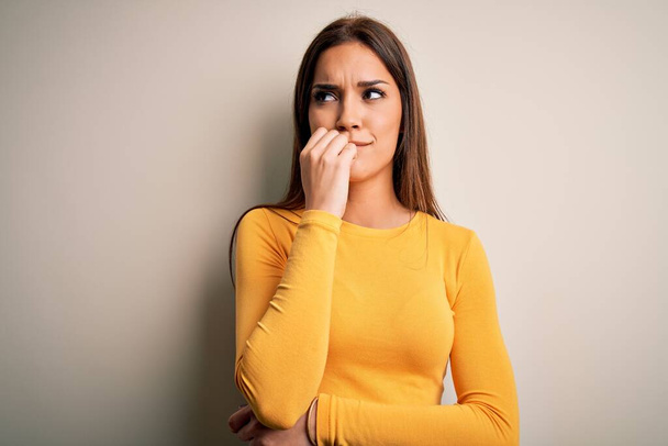Young beautiful brunette woman wearing yellow casual t-shirt over white background looking stressed and nervous with hands on mouth biting nails. Anxiety problem. - Фото, изображение