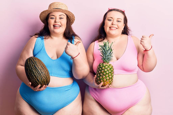 Dos jovencitos más grandes que llevan bikini sosteniendo melón y piña apuntando pulgar hasta el lado sonriendo felices con la boca abierta.  - Foto, Imagen