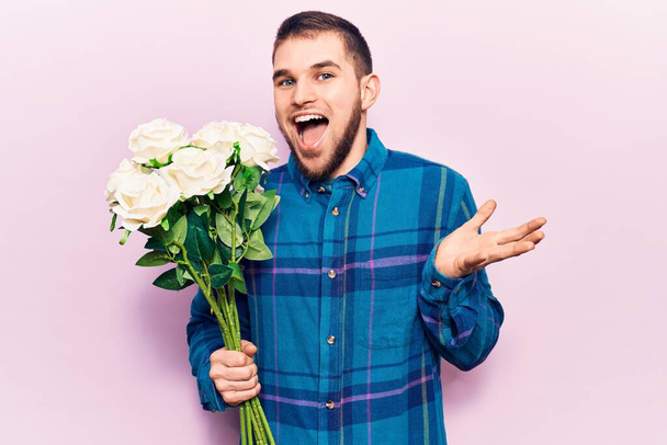 Νεαρός όμορφος άνδρας κρατώντας μπουκέτο λουλούδια γιορτάζει το επίτευγμα με χαρούμενο χαμόγελο και την έκφραση του νικητή με υψωμένο χέρι  - Φωτογραφία, εικόνα