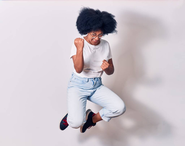 Молода афро-американська жінка, одягнена в повсякденний одяг і окуляри, посміхаються, щасливі. Стрибаючи з посмішкою на обличчі роблячи жест переможця з кулаками над ізольованим білим фоном - Фото, зображення