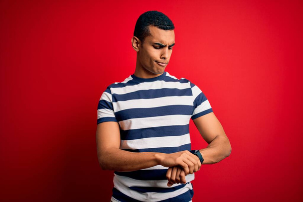 Bonito homem afro-americano vestindo camiseta listrada casual em pé sobre fundo vermelho Verificando o tempo no relógio de pulso, relaxado e confiante
 - Foto, Imagem