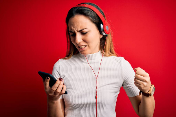 Young beautiful brunette woman listening to music using smartphone and headphones annoyed and frustrated shouting with anger, crazy and yelling with raised hand, anger concept - Фото, изображение